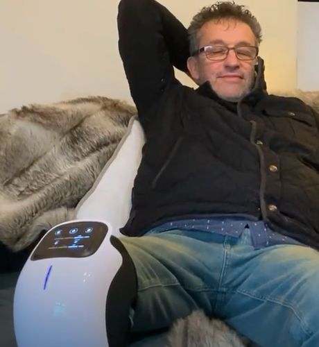 Man sitting on a white robot-like device with a remote control, wearing a black jacket and blue jeans.